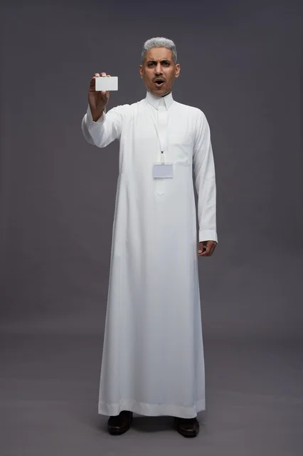 بورتريه لرجل عربي سعودي خليجي يرتدي الثوب الابيض يعرض بطاقة ائتمان بيضاء فارغه ويشير اليها, كرت تعريفي شخصي, الجسم بالكامل, خلفيه رماديه