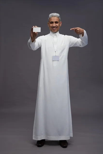 A portrait of an Arab Saudi Gulf man wearing a white thobe displaying a blank white credit card and pointing to it, a personal identification card, full body, gray background.