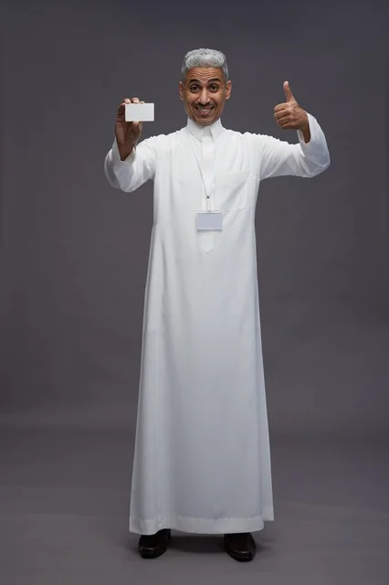 بورتريه لرجل عربي سعودي خليجي يرتدي الثوب الابيض يعرض بطاقة ائتمان بيضاء فارغه ويشير اليها, كرت تعريفي شخصي, الجسم بالكامل, خلفيه رماديه