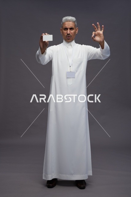 بورتريه لرجل عربي سعودي خليجي يرتدي الثوب الابيض يعرض بطاقة ائتمان بيضاء فارغه ويشير اليها, كرت تعريفي شخصي, الجسم بالكامل, خلفيه رماديه