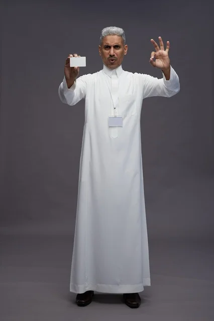بورتريه لرجل عربي سعودي خليجي يرتدي الثوب الابيض يعرض بطاقة ائتمان بيضاء فارغه ويشير اليها, كرت تعريفي شخصي, الجسم بالكامل, خلفيه رماديه