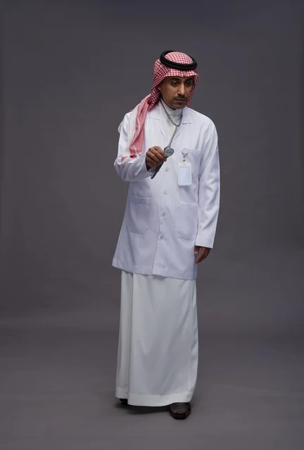 طبيب عربي سعودي خليجي يرتدي الثوب والمعطف الابيض مع سماعة الطب يقوم بايماءات واشارات مختلفه, كامل الجسم, خلفيه رماديه