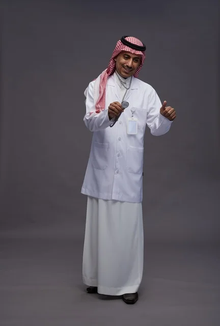 طبيب عربي سعودي خليجي يرتدي الثوب والمعطف الابيض مع سماعة الطب يقوم بايماءات واشارات مختلفه, كامل الجسم, خلفيه رماديه