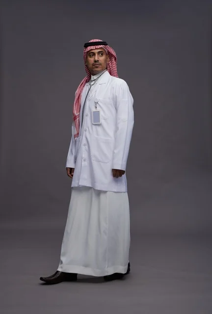 طبيب عربي سعودي خليجي يرتدي الثوب والمعطف الابيض مع سماعة الطب يقوم بايماءات واشارات مختلفه, كامل الجسم, خلفيه رماديه