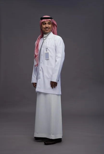 طبيب عربي سعودي خليجي يرتدي الثوب والمعطف الابيض مع سماعة الطب يقوم بايماءات واشارات مختلفه, كامل الجسم, خلفيه رماديه