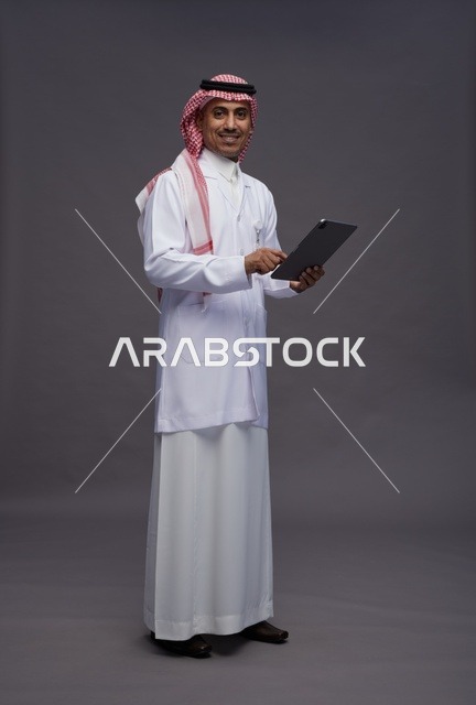 طبيب عربي سعودي خليجي يرتدي الثوب والمعطف الابيض  يحمل بيده جهاز لوحي, خدمات الرعايه الصحيه الرقميه, كامل الجسم, خلفيه رماديه