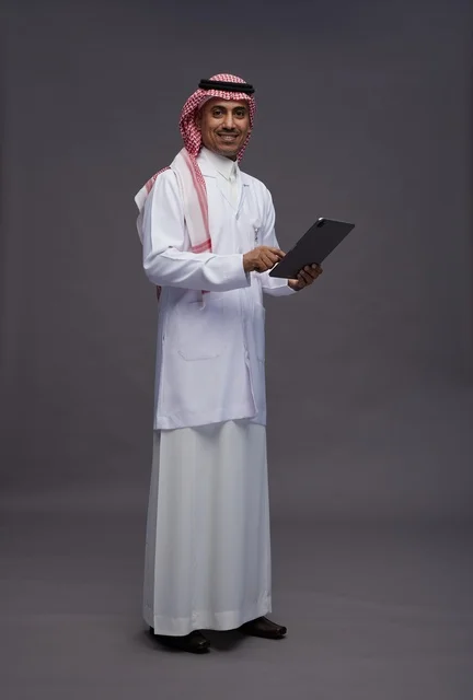 طبيب عربي سعودي خليجي يرتدي الثوب والمعطف الابيض  يحمل بيده جهاز لوحي, خدمات الرعايه الصحيه الرقميه, كامل الجسم, خلفيه رماديه
