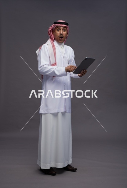 طبيب عربي سعودي خليجي يرتدي الثوب والمعطف الابيض  يحمل بيده جهاز لوحي, خدمات الرعايه الصحيه الرقميه, كامل الجسم, خلفيه رماديه