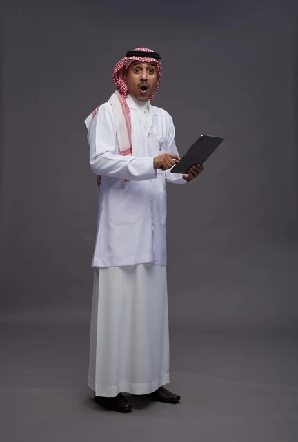 طبيب عربي سعودي خليجي يرتدي الثوب والمعطف الابيض  يحمل بيده جهاز لوحي, خدمات الرعايه الصحيه الرقميه, كامل الجسم, خلفيه رماديه