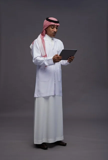 طبيب عربي سعودي خليجي يرتدي الثوب والمعطف الابيض  يحمل بيده جهاز لوحي, خدمات الرعايه الصحيه الرقميه, كامل الجسم, خلفيه رماديه