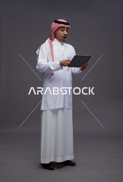 طبيب عربي سعودي خليجي يرتدي الثوب والمعطف الابيض  يحمل بيده جهاز لوحي, خدمات الرعايه الصحيه الرقميه, كامل الجسم, خلفيه رماديه
