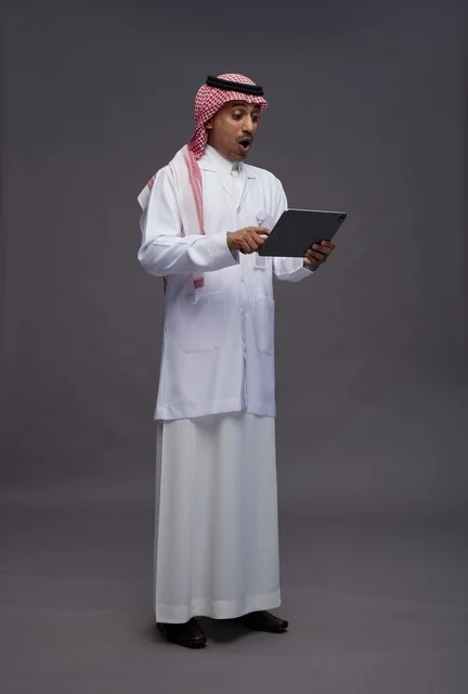 طبيب عربي سعودي خليجي يرتدي الثوب والمعطف الابيض  يحمل بيده جهاز لوحي, خدمات الرعايه الصحيه الرقميه, كامل الجسم, خلفيه رماديه