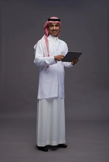 طبيب عربي سعودي خليجي يرتدي الثوب والمعطف الابيض  يحمل بيده جهاز لوحي, خدمات الرعايه الصحيه الرقميه, كامل الجسم, خلفيه رماديه