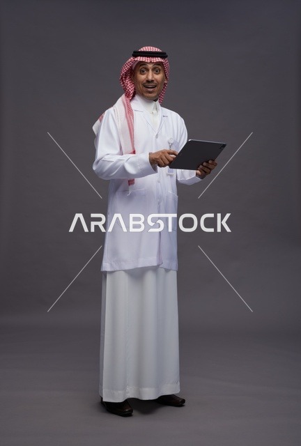 طبيب عربي سعودي خليجي يرتدي الثوب والمعطف الابيض  يحمل بيده جهاز لوحي, خدمات الرعايه الصحيه الرقميه, كامل الجسم, خلفيه رماديه
