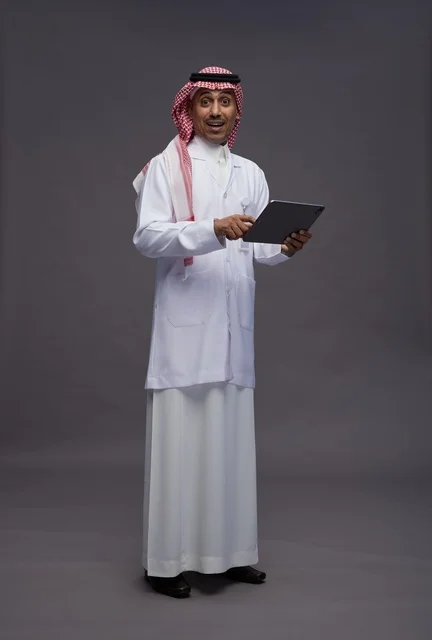 An Arab Saudi Gulf doctor wearing a thobe and a white coat holding a tablet in his hand, digital healthcare services, full body, gray background.