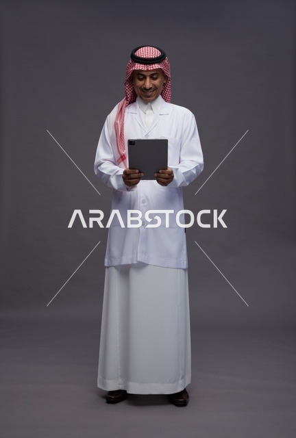 طبيب عربي سعودي خليجي يرتدي الثوب والمعطف الابيض  يحمل بيده جهاز لوحي, خدمات الرعايه الصحيه الرقميه, كامل الجسم, خلفيه رماديه