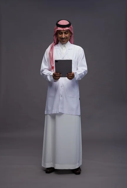 طبيب عربي سعودي خليجي يرتدي الثوب والمعطف الابيض  يحمل بيده جهاز لوحي, خدمات الرعايه الصحيه الرقميه, كامل الجسم, خلفيه رماديه