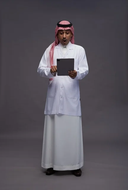 طبيب عربي سعودي خليجي يرتدي الثوب والمعطف الابيض  يحمل بيده جهاز لوحي, خدمات الرعايه الصحيه الرقميه, كامل الجسم, خلفيه رماديه