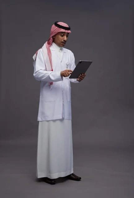 طبيب عربي سعودي خليجي يرتدي الثوب والمعطف الابيض  يحمل بيده جهاز لوحي, خدمات الرعايه الصحيه الرقميه, كامل الجسم, خلفيه رماديه