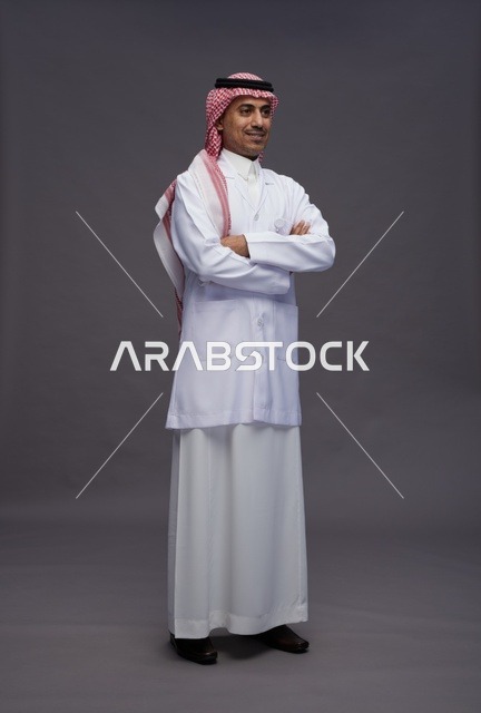 طبيب عربي سعودي خليجي يرتدي الثوب والمعطف الابيض متكتف اليدين, الوقوف بثقه بايدي متقاطعه, كامل الجسم, خلفيه رماديه