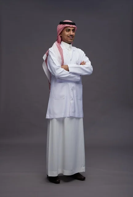 A Saudi Arabian Gulf Arab doctor wearing a thobe and a white coat, standing confidently with arms crossed, full body, gray background.