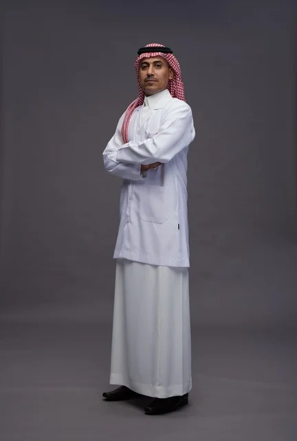 A Saudi Arabian Gulf Arab doctor wearing a thobe and a white coat, standing confidently with arms crossed, full body, gray background.