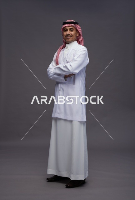 طبيب عربي سعودي خليجي يرتدي الثوب والمعطف الابيض متكتف اليدين, الوقوف بثقه بايدي متقاطعه, كامل الجسم, خلفيه رماديه