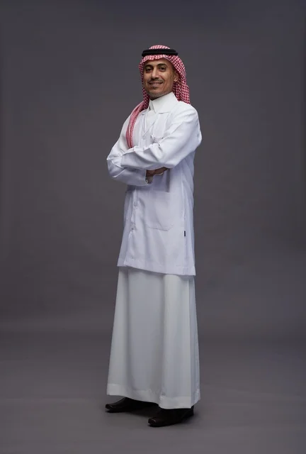 طبيب عربي سعودي خليجي يرتدي الثوب والمعطف الابيض متكتف اليدين, الوقوف بثقه بايدي متقاطعه, كامل الجسم, خلفيه رماديه