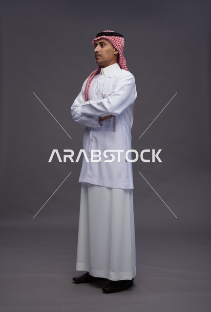 طبيب عربي سعودي خليجي يرتدي الثوب والمعطف الابيض متكتف اليدين, الوقوف بثقه بايدي متقاطعه, كامل الجسم, خلفيه رماديه