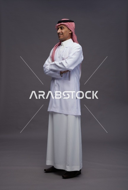 طبيب عربي سعودي خليجي يرتدي الثوب والمعطف الابيض متكتف اليدين, الوقوف بثقه بايدي متقاطعه, كامل الجسم, خلفيه رماديه