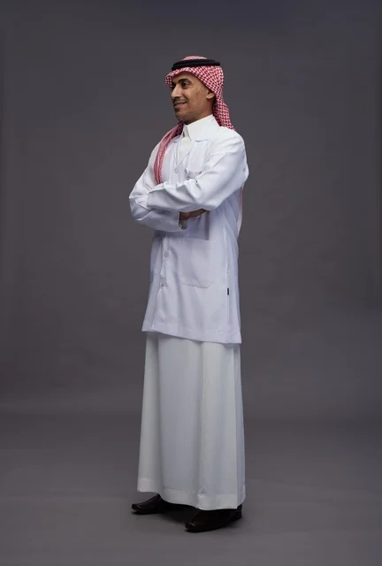 A Saudi Arabian Gulf Arab doctor wearing a thobe and a white coat, standing confidently with arms crossed, full body, gray background.