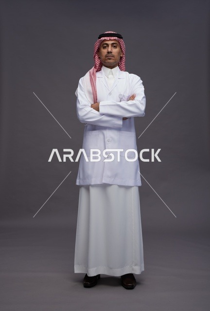 طبيب عربي سعودي خليجي يرتدي الثوب والمعطف الابيض متكتف اليدين, الوقوف بثقه بايدي متقاطعه, كامل الجسم, خلفيه رماديه