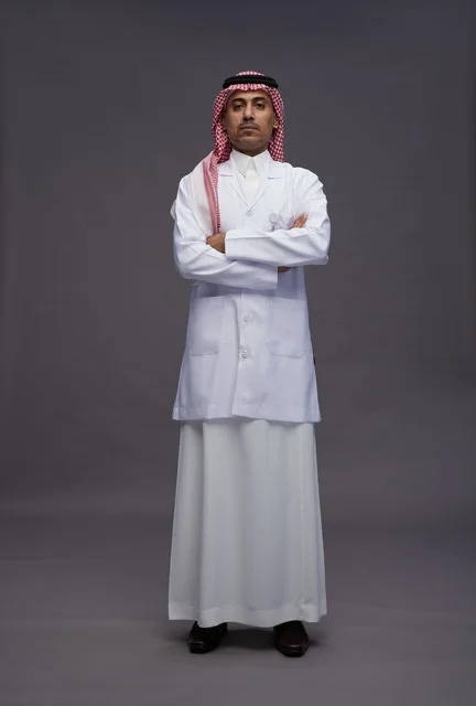 A Saudi Arabian Gulf Arab doctor wearing a thobe and a white coat, standing confidently with arms crossed, full body, gray background.