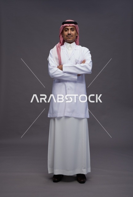 طبيب عربي سعودي خليجي يرتدي الثوب والمعطف الابيض متكتف اليدين, الوقوف بثقه بايدي متقاطعه, كامل الجسم, خلفيه رماديه