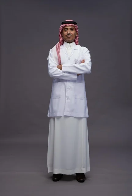 A Saudi Arabian Gulf Arab doctor wearing a thobe and a white coat, standing confidently with arms crossed, full body, gray background.