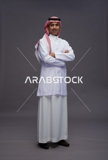 طبيب عربي سعودي خليجي يرتدي الثوب والمعطف الابيض متكتف اليدين, الوقوف بثقه بايدي متقاطعه, كامل الجسم, خلفيه رماديه