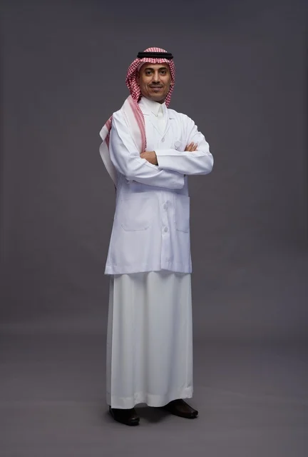A Saudi Arabian Gulf Arab doctor wearing a thobe and a white coat, standing confidently with arms crossed, full body, gray background.