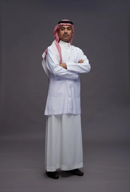 A Saudi Arabian Gulf Arab doctor wearing a thobe and a white coat, standing confidently with arms crossed, full body, gray background.