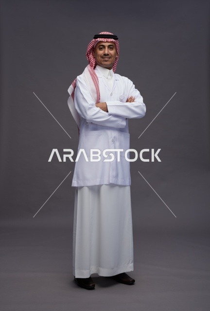طبيب عربي سعودي خليجي يرتدي الثوب والمعطف الابيض متكتف اليدين, الوقوف بثقه بايدي متقاطعه, كامل الجسم, خلفيه رماديه