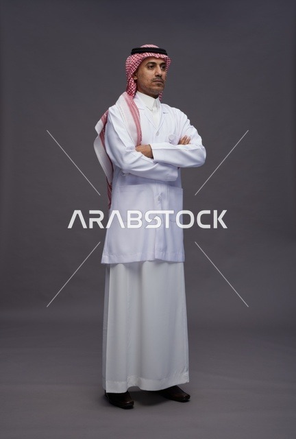 طبيب عربي سعودي خليجي يرتدي الثوب والمعطف الابيض متكتف اليدين, الوقوف بثقه بايدي متقاطعه, كامل الجسم, خلفيه رماديه