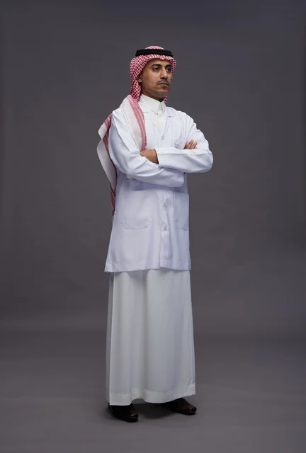طبيب عربي سعودي خليجي يرتدي الثوب والمعطف الابيض متكتف اليدين, الوقوف بثقه بايدي متقاطعه, كامل الجسم, خلفيه رماديه
