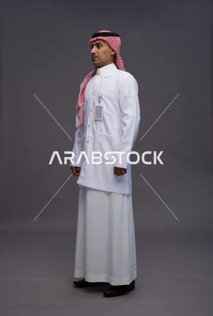 طبيب عربي سعودي خليجي يرتدي الثوب والمعطف الابيض ينظر الى الكاميرا, الوقوف بثقه , كامل الجسم, خلفيه رماديه