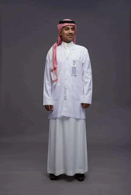 طبيب عربي سعودي خليجي يرتدي الثوب والمعطف الابيض ينظر الى الكاميرا, الوقوف بثقه , كامل الجسم, خلفيه رماديه