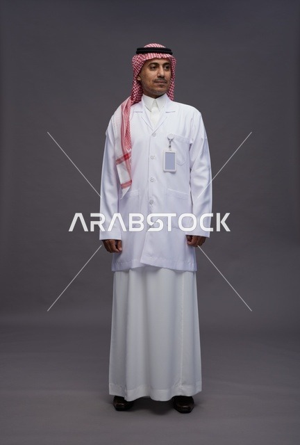 طبيب عربي سعودي خليجي يرتدي الثوب والمعطف الابيض ينظر الى الكاميرا, الوقوف بثقه , كامل الجسم, خلفيه رماديه