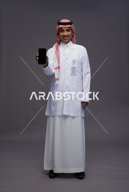 طبيب عربي سعودي خليجي يرتدي الثوب والشماغ مع المعطف الطبي يستخدم الهاتف المحمول, الخدمات الصحيه الرقميه, كامل الجسم, خلفيه رماديه