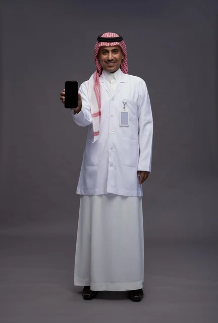 طبيب عربي سعودي خليجي يرتدي الثوب والشماغ مع المعطف الطبي يستخدم الهاتف المحمول, الخدمات الصحيه الرقميه, كامل الجسم, خلفيه رماديه