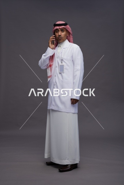 طبيب عربي سعودي خليجي يرتدي الثوب والشماغ مع المعطف الطبي يستخدم الهاتف المحمول, الخدمات الصحيه الرقميه, كامل الجسم, خلفيه رماديه
