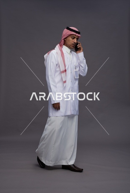 طبيب عربي سعودي خليجي يرتدي الثوب والشماغ مع المعطف الطبي يستخدم الهاتف المحمول, الخدمات الصحيه الرقميه, كامل الجسم, خلفيه رماديه