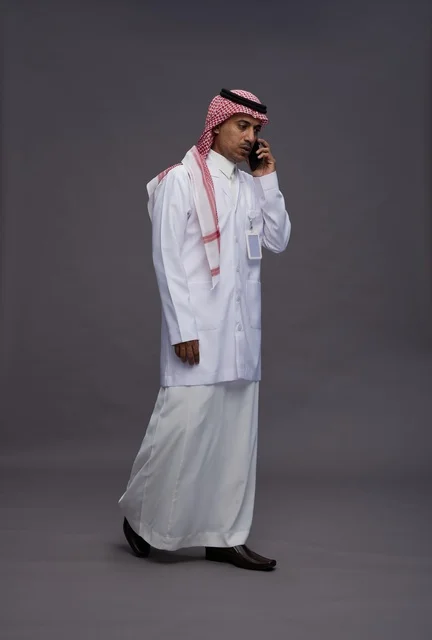 طبيب عربي سعودي خليجي يرتدي الثوب والشماغ مع المعطف الطبي يستخدم الهاتف المحمول, الخدمات الصحيه الرقميه, كامل الجسم, خلفيه رماديه