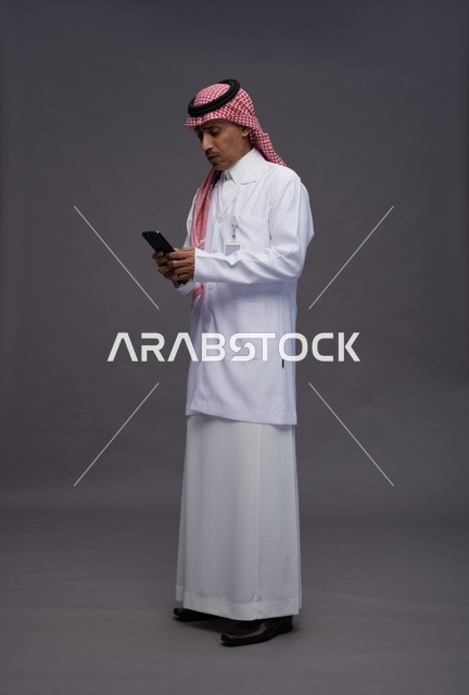 A Saudi Arabian Gulf Arab doctor wearing a thobe and shemagh with a medical coat using a mobile phone, digital health services, full body, gray background.